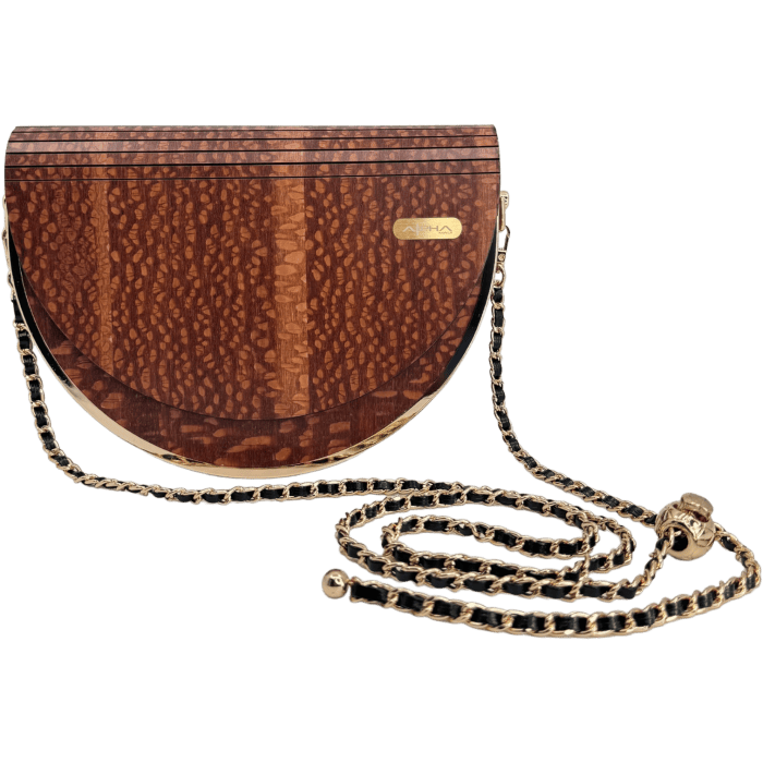 The Alpha Maui Pu'olo Handbag is a half-circle wooden accessory adorned with a textured brown surface and a small gold emblem on the upper right corner. It boasts a long, gold chain strap interwoven with black thread, making it an exquisite addition to your beach attire in Hawaii. Its sleek and elegant design encapsulates the essence of Alpha Maui sophistication.