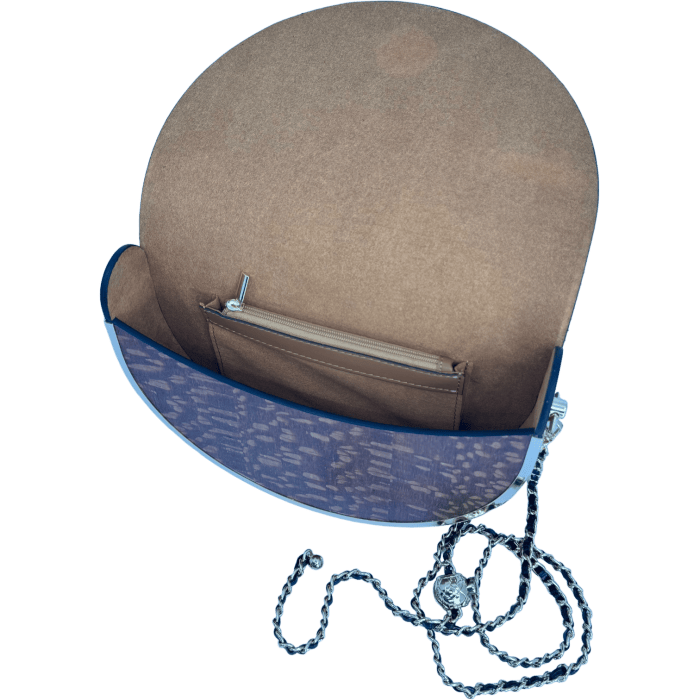 The Alpha Maui Pu'olo Handbag, featuring a blue exterior with a round flap, is open to display its brown suede interior and zippered pocket. Beside the bag lies a coiled silver chain strap. The outer fabric, inspired by Alpha Maui designs, showcases a subtle pattern.