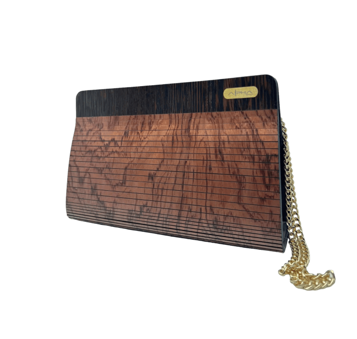The Alpha Maui The Clutch Carrier is a rectangular clutch purse adorned with a wooden texture design on the front, showcasing dark and light brown tones arranged in horizontal lines. Accompanied by a gold chain strap elegantly draped beside it, the clutch proudly displays the brand logo in the top right corner, evoking the Aloha spirit. The background is sleek black.