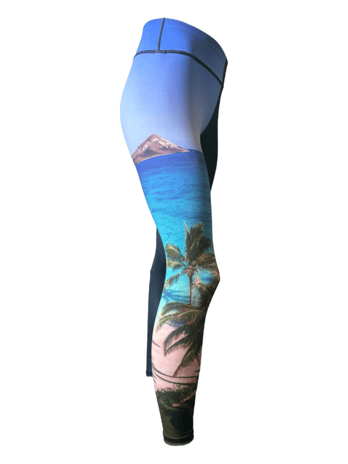 A side view of the Alpha Maui Waimanalo (Full Length) leggings showcases a tropical beach scene adorned with palm trees, white sand, turquoise water, and a distant mountain under a clear blue sky. The design wraps around the lower leg, creating an Aloha-inspired, vibrant and summery look.