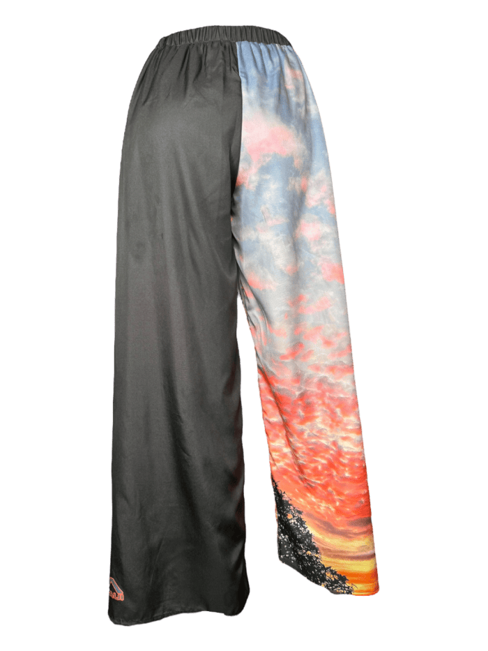 Introducing the Alpha Maui Pukalani Wrap Pants: wide-legged pants with a unique split design. The left side is solid black, while the right side features a vibrant sunset scene with hues of orange, pink, and blue, accompanied by tree silhouettes at the bottom. Channeling Alpha Maui's Hawaii vibe, these pants embody the Aloha spirit with an elastic waistband for added comfort.