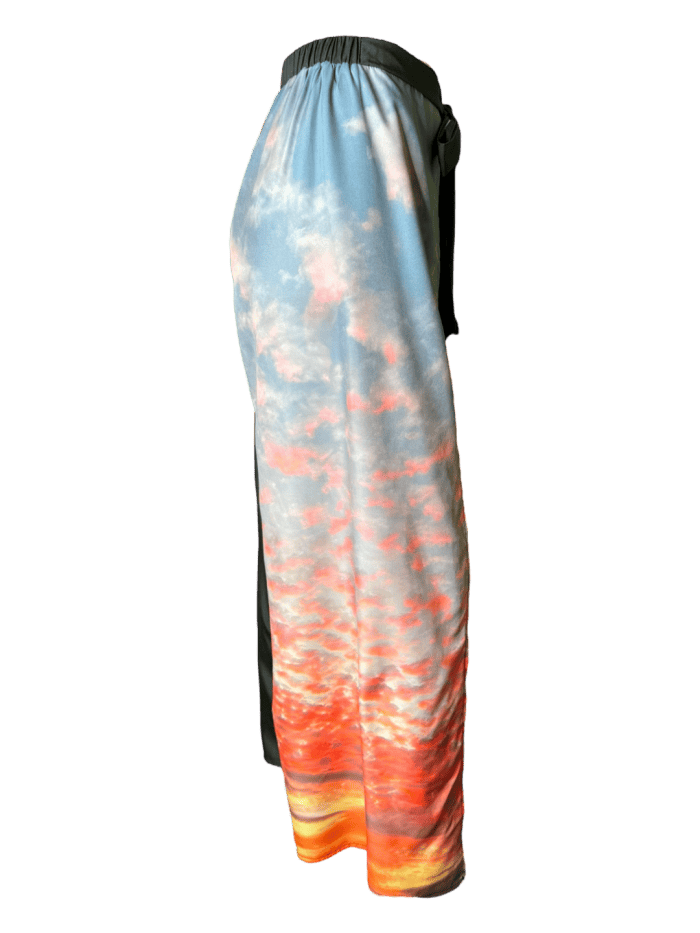 A side view of the Alpha Maui Pukalani Wrap Pants showcasing an evening sky print adorned with soft, multicolored clouds in shades of pink, orange, and yellow. The waistband is black, and the material boasts a smooth, flowing texture. These wrap pants drape elegantly, highlighting the vivid, picturesque design reminiscent of a Hawaiian sunset.