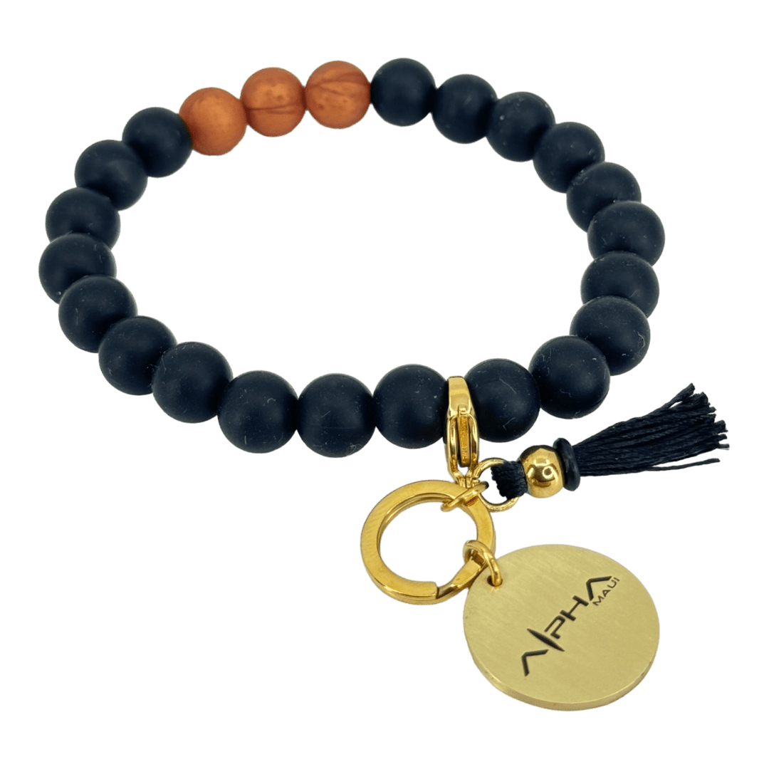 The Alpha Silicone Bracelet is predominantly black beads punctuated by three brown beads. It includes a key ring adorned with a round, gold-colored tag etched with "APHA" and a small tassel. Evoking Hawaii’s serene beaches, this bracelet blends matte and polished textures, making it an ideal addition to your beach apparel collection.