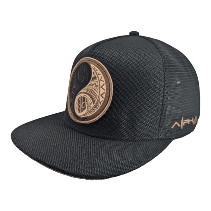 The Balance is a black mesh baseball cap adorned with an intricate brown circular design on the front. This design features detailed patterns and shapes, and the "ALPHA" logo is embroidered in brown on the side. The cap includes a structured front panel and a curved brim.