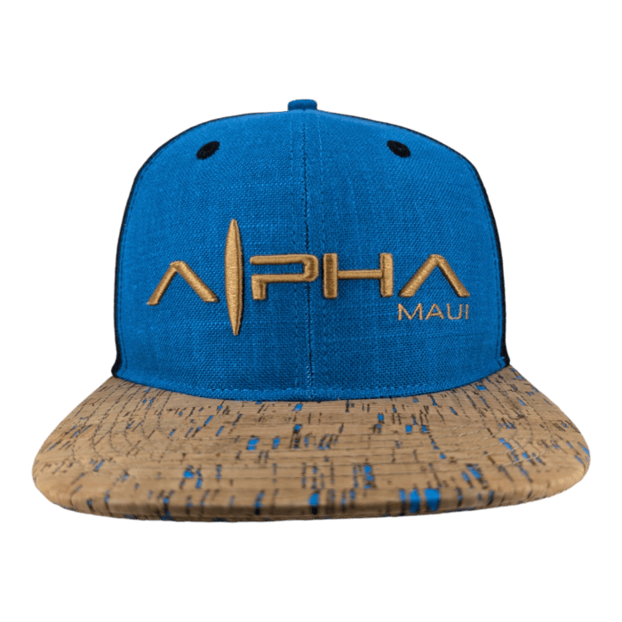 A blue snapback cap named "The Coastline" features the text "ALPHA" in yellow letters along with "MAUI" in smaller yellow letters to the right. Its brim has a cork-like appearance with blue speckles, and it includes two small, black ventilation holes on the front panel.