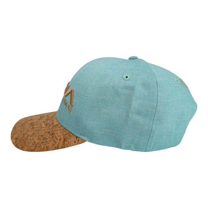 The Turtle Cove II baseball cap, in a striking teal color, is displayed with its unique cork material brim. The cap is adorned with an embroidered yellow logo on the side. A black backdrop accentuates the vibrant colors and textures of the hat.