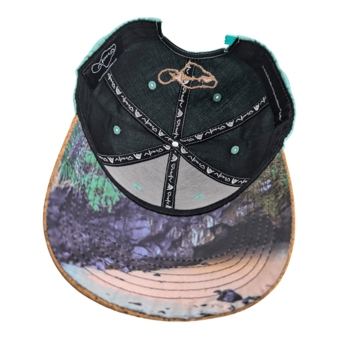 The image displays the inside of the Turtle Cove II baseball cap, featuring a vibrant, patterned interior. The sweatband and seams are crafted from black fabric with teal stitching and adorned with the brand's logo. The underside of the visor showcases a detailed, colorful graphic of a natural landscape.