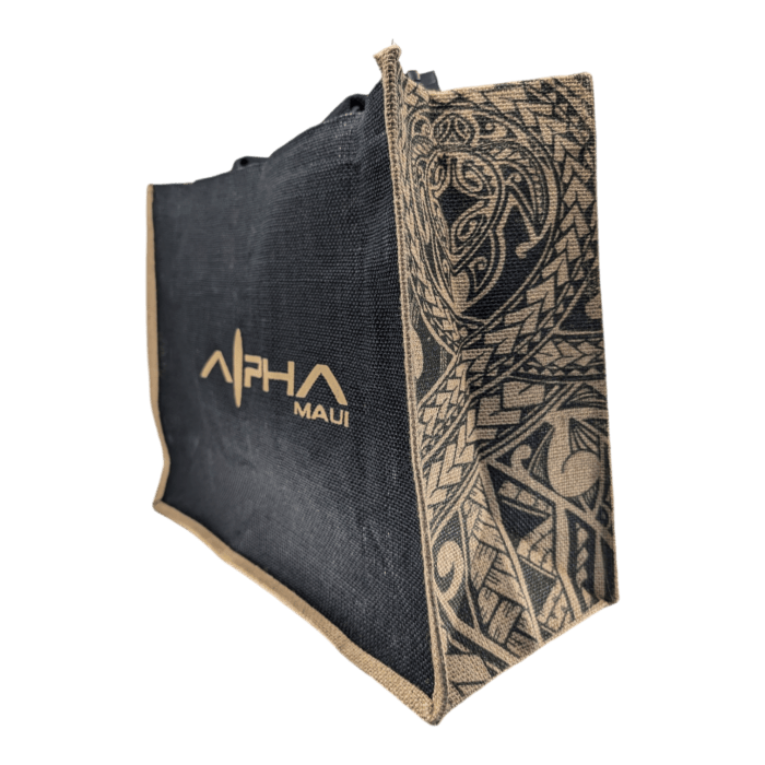 The Tribal Wai tote bag combines black and natural colors with intricate tribal-style designs on the side panels. The front features "Alpha Maui" in gold lettering, and it's made from textured, woven fabric for a durable and stylish look.