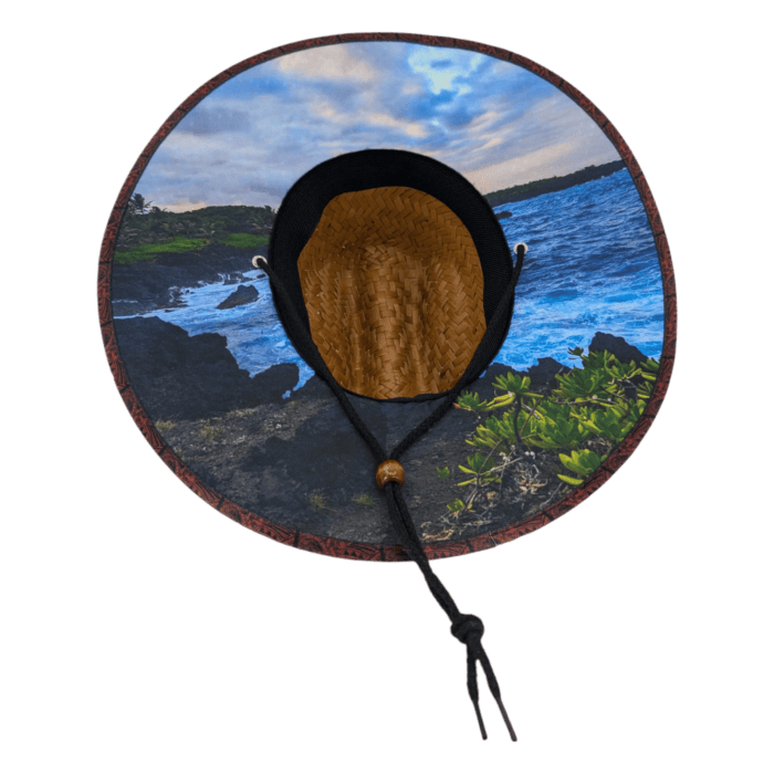 The Autumn Sunrise features a wide-brimmed straw hat with a scenic landscape printed on the brim's underside, showcasing a coastal view of rocky cliffs, green foliage, and ocean waves beneath a partly cloudy sky. The hat is accented with a black strap and wooden bead.