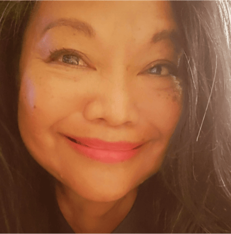 ADELAIDE-SHERMAN A person with long dark hair smiles warmly at the camera, with soft lighting highlighting their features. They are wearing makeup, including pink lipstick, and have a gentle, cheerful expression.