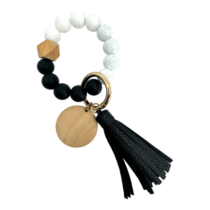 A bracelet with alternating black, white, and speckled beads, featuring a wooden hexagonal bead, a gold keyring, a round wooden disc, and a black tassel.
