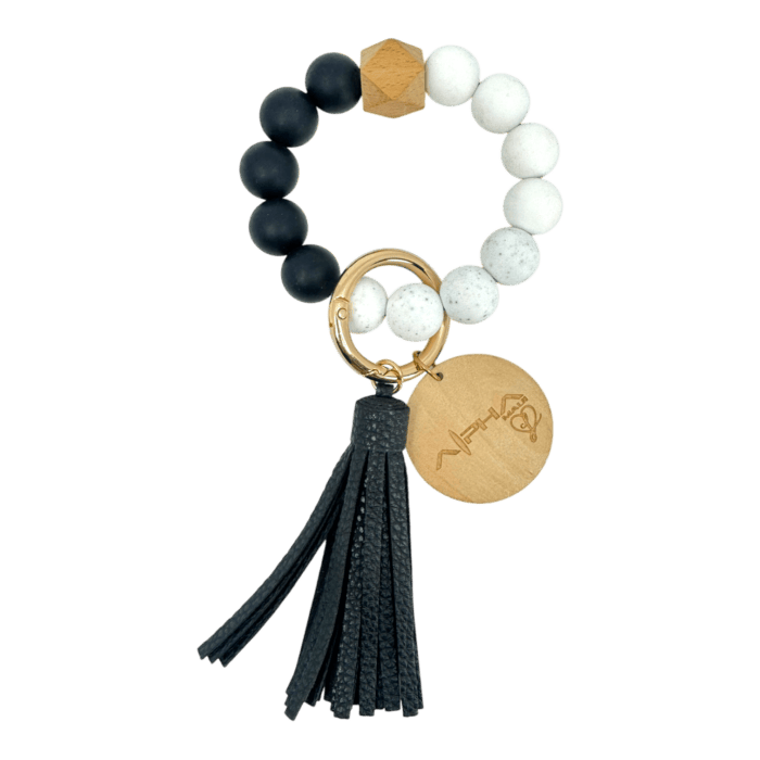 A stylish keychain featuring a bracelet made of alternating black and white beads, a wooden hexagonal bead, and a gold ring attached to a black tassel and wooden charm with an engraved design.