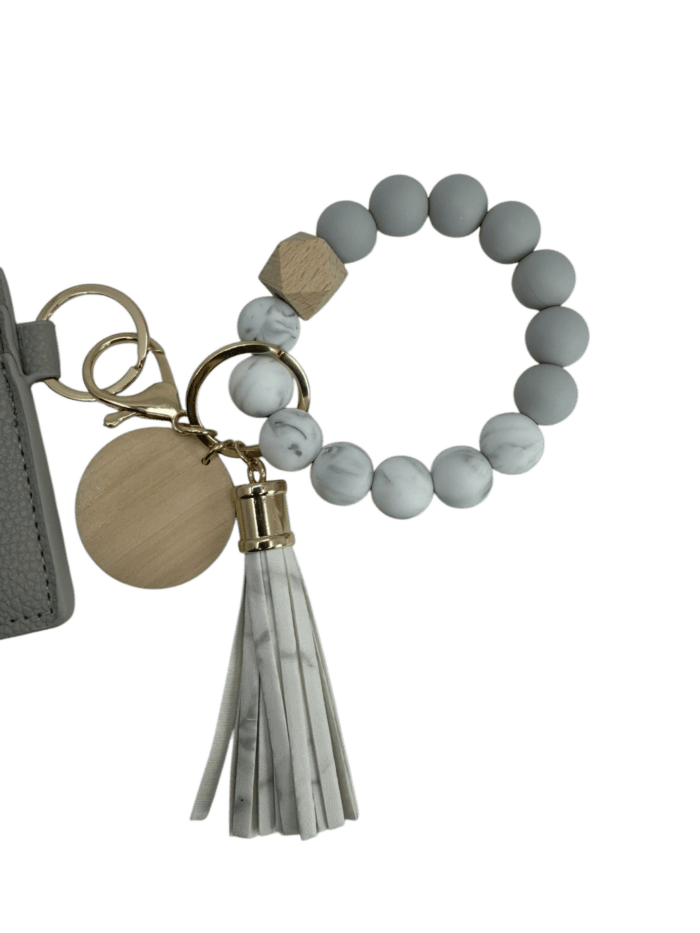A keychain featuring a round wooden disc, a gray beaded bracelet, and a white leather tassel. The beads have a marbled design, and a wooden hexagonal bead adds an accent. The keychain is attached to a textured gray strap.