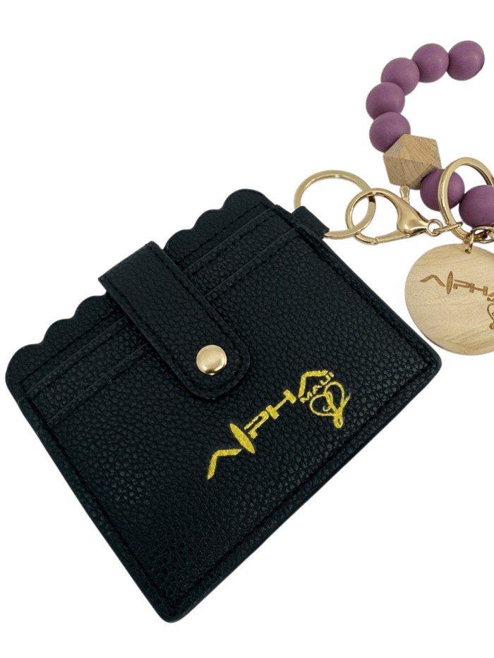A black leather cardholder with a scalloped edge and snap button, featuring a gold logo. Attached is a keychain with purple beads and a round wooden tag engraved with a design.